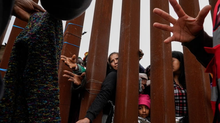 Migrants reach through a border wall for clothing handed out...