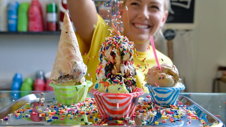 An ice cream server adds rainbow sprinkles to three scoops...