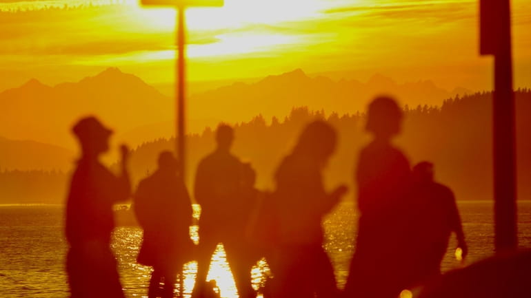The sun sets behind the Olympic Mountains as people cross...
